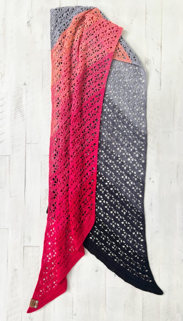 A crochet bias shawl showcasing a stunning gradient yarn draped over a white wooden surface. The colors transition smoothly from light gray at the top to deep pink and red at the bottom, featuring an intricate lace design. -Marly Bird