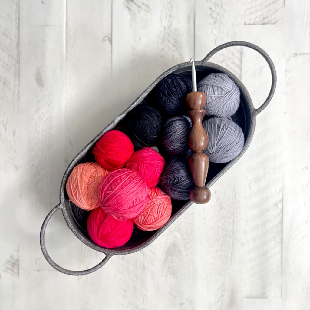 A metal basket filled with various colored yarn balls including shades of pink, red, black, and gray. A wooden-handled crochet hook rests on top. The basket is on a white wooden surface. -Marly Bird