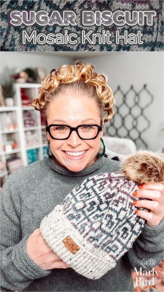 A smiling woman with glasses and curly hair holds up a knit hat with a fur pom-pom, featuring a colorful mosaic pattern. Text above reads: "Sugar Biscuit Knit Hat Pattern." The Marly Bird logo is in the corner. -Marly Bird