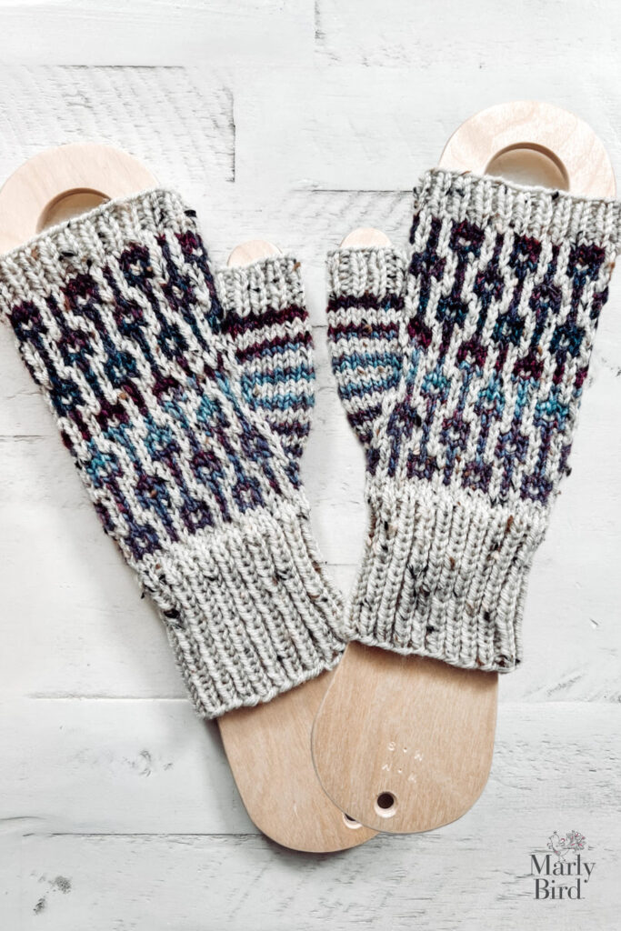 Two knitted fingerless gloves with colorful mosaic patterns in shades of blue, purple, and gray on light wooden sock blockers. Featuring a vibrant Marly Bird design, these Knit Fingerless Mitts Pattern are set on a white wooden background. -Marly Bird