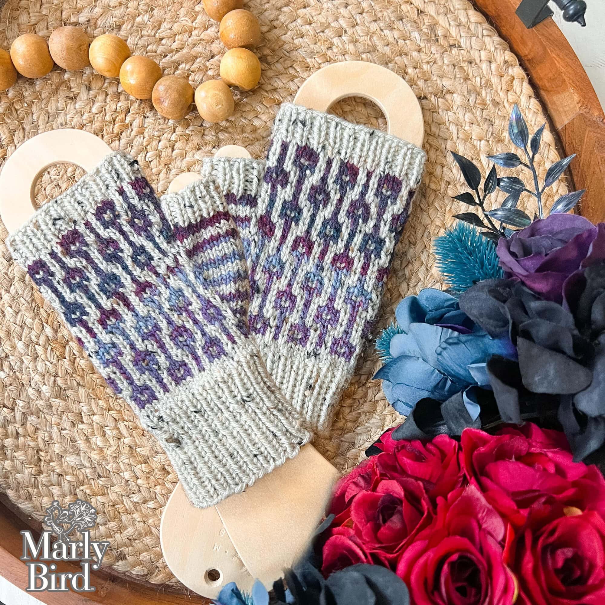 Two knitted fingerless gloves in the Sugar Biscuit colorway rest on wooden inserts, placed on a woven tray with beads and artificial blue and red flowers. "Marly Bird" is written in the bottom left corner, showcasing this Knit Fingerless Mitts Pattern. -Marly Bird