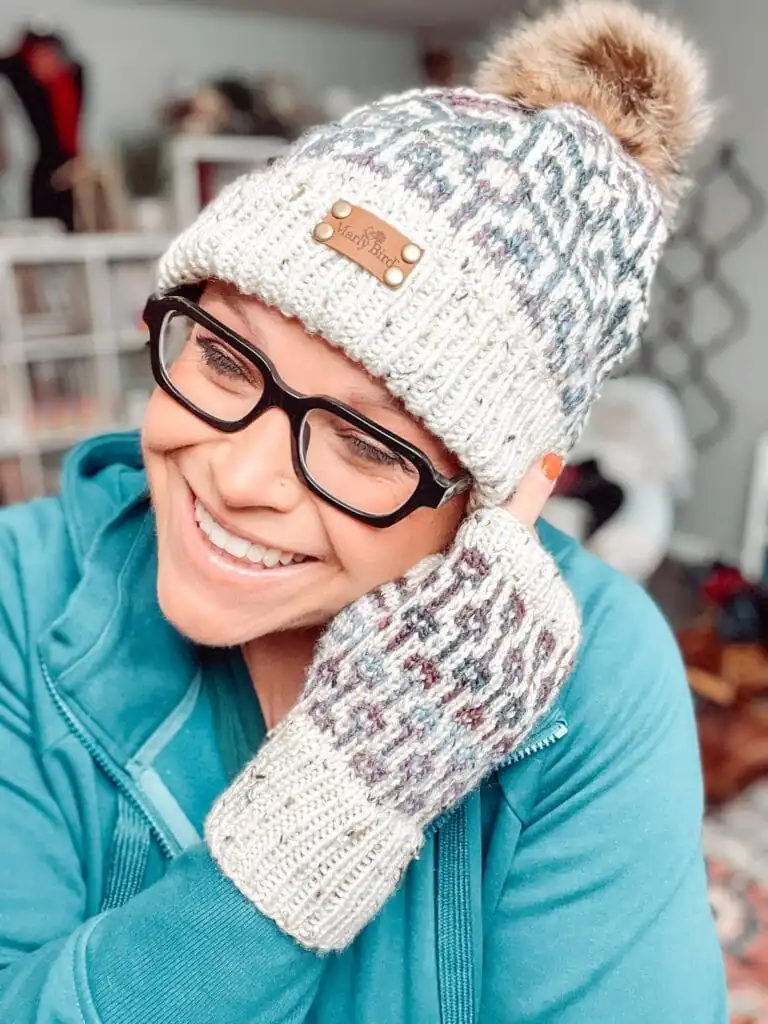 Smiling person wearing black glasses, teal hoodie, Marly Bird knit hat with a pom-pom, and matching Sugar Biscuit Knit Fingerless Mitts Pattern mittens, touching their face in a cozy indoor setting with shelves and blurred background. -Marly Bird