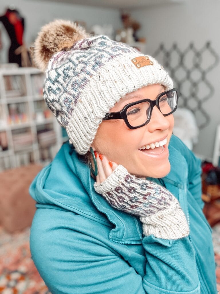 A smiling person wearing glasses, a knit hat with a pom-pom, and matching fingerless mitts made from the Sugar Biscuit Knit pattern by Marly Bird, poses indoors with bookshelves and decor in the background. -Marly Bird