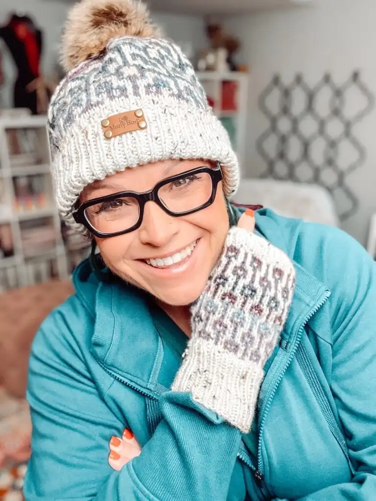 A smiling person wearing black glasses, a Marly Bird patterned knit hat with a pom-pom, and matching knit mittens poses indoors, resting their gloved hand on their chin. They are dressed in a teal zip-up jacket. -Marly Bird
