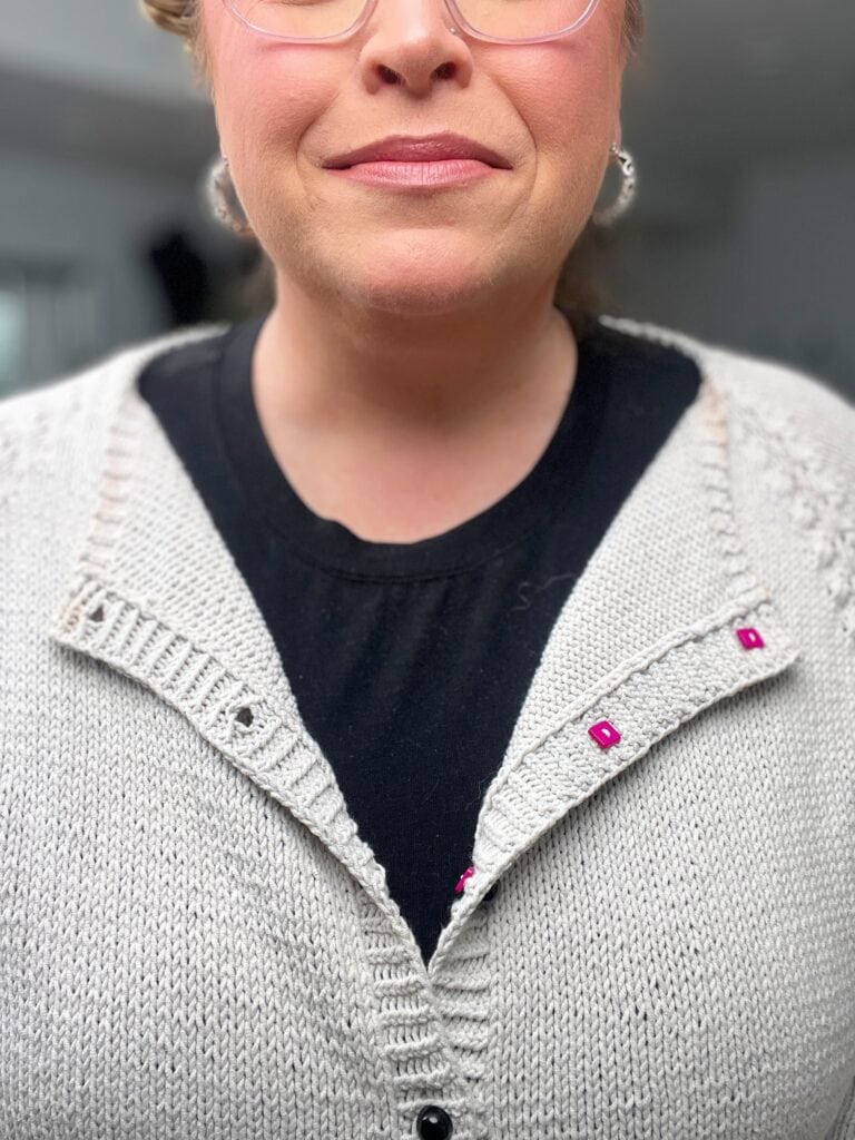 A person wearing a light gray knitted cardigan over a black shirt. The sugar frost knit cardigan’s collar is held together with small pink stitch markers. The person's lower face and upper chest are visible. -Marly Bird