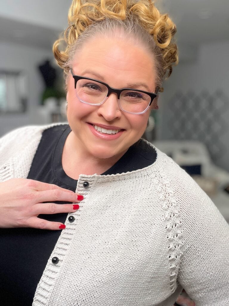 A smiling person with curly blonde hair, glasses, and red nail polish wears a light Marly Bird knit cardigan over a black top, posing indoors and looking at the camera. -Marly Bird