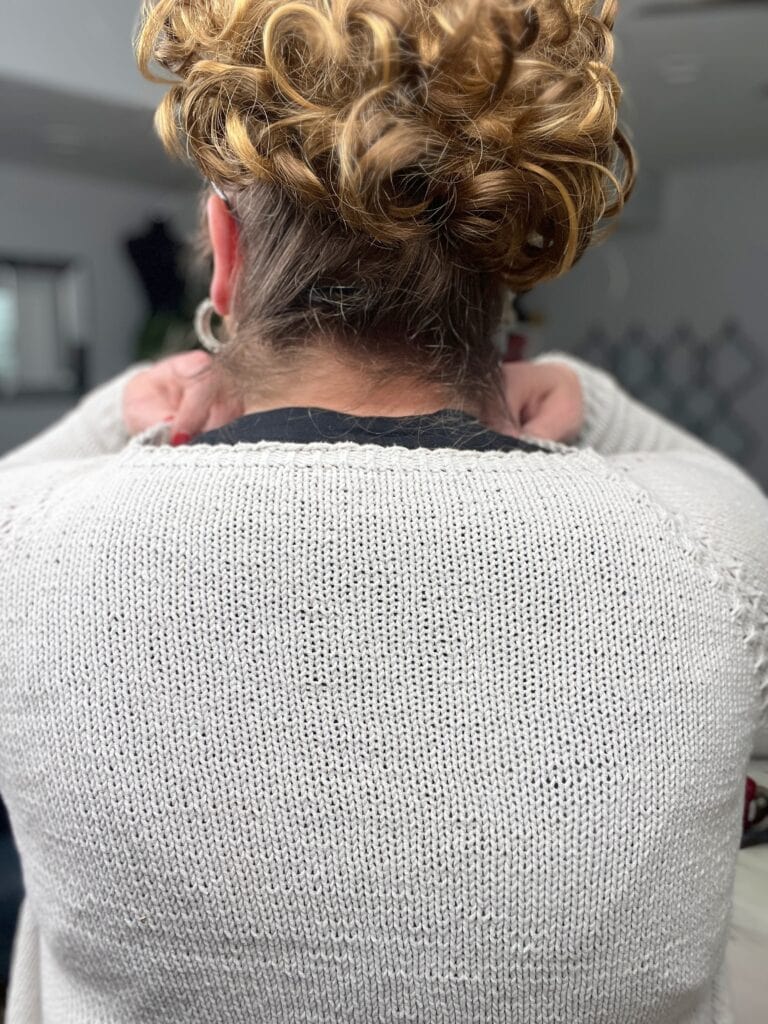 A person with curly, light brown hair tied up is seen from behind, wearing a light gray knit cardigan pattern inspired by Marly Bird and holding their hands near their neck. The background is indoors and slightly blurred. -Marly Bird
