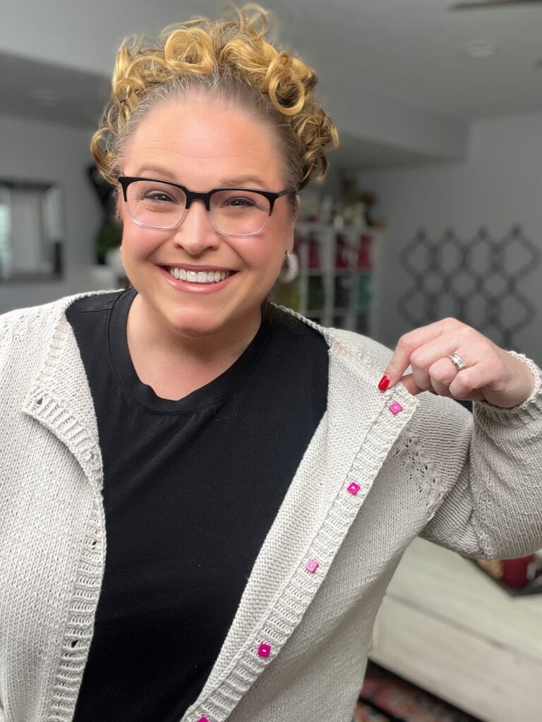 A smiling woman with curly hair and glasses shows off pink snap buttons on the inside of her light knit cardigan, pulling it open to display the buttons. She is wearing a black shirt underneath—perfect inspiration for your next Marly Bird project!. -Marly Bird