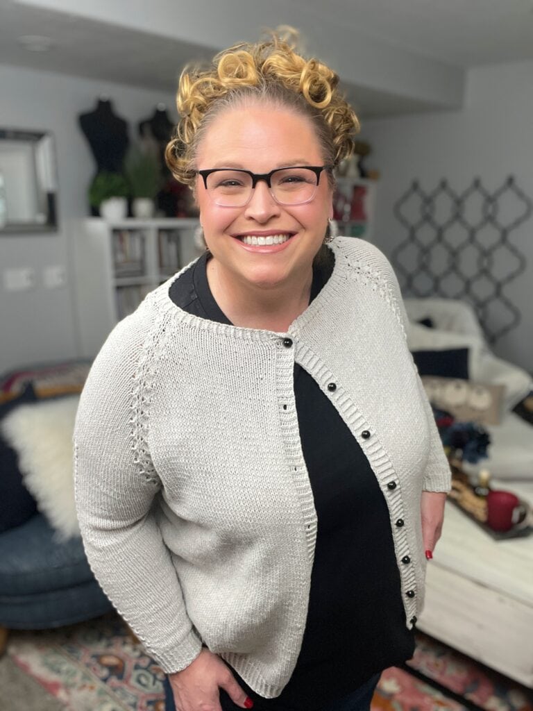 A smiling woman with curly blonde hair and glasses wears a light-colored knit cardigan, inspired by a Marly Bird Knit Cardigan Pattern, over a black top, standing in a cozy, well-decorated living room. -Marly Bird