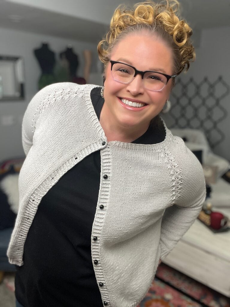 A smiling person with glasses and curly hair wears a light-colored knit cardigan over a black shirt, standing indoors in a cozy room with home decor, sewing mannequins, and hints of a Free Knit Cardigan Pattern by Marly Bird in the background. -Marly Bird