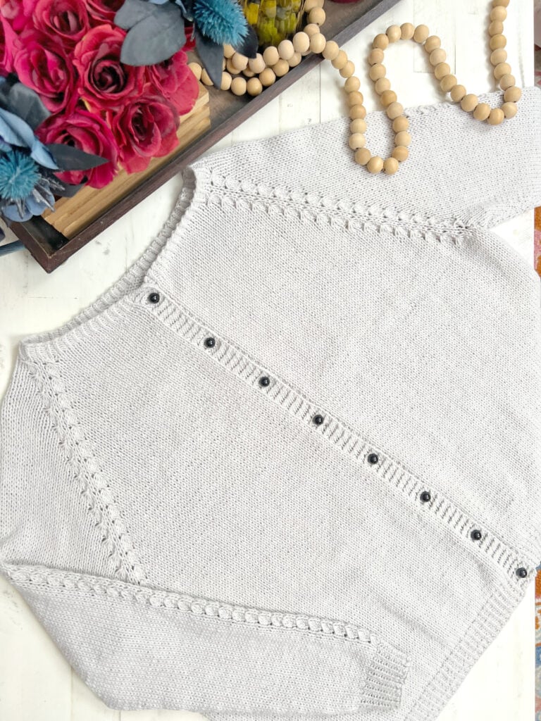 A light gray Sugar Frost knit cardigan with black buttons is laid out flat on a white surface next to a beaded necklace and a wooden tray with pink and blue flowers. The cardigan features a decorative textured pattern on the sleeves and edges. -Marly Bird