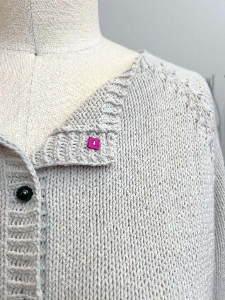 A close-up of a cream-colored knitted cardigan on a dress form, featuring a pink square stitch marker near the neckline and black buttons down the front—a lovely Free Knit Cardigan pattern inspired by Marly Bird. -Marly Bird