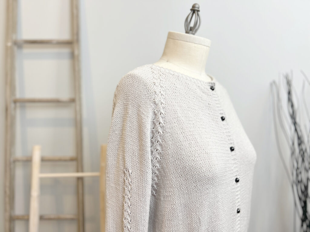 A mannequin displays the Sugar Frost Knit Cardigan, a light gray knitted sweater with black buttons and lace detailing on the sleeves, set against a blurred background with a wooden ladder and decorative branches. -Marly Bird