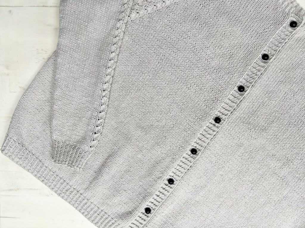 A light gray Sugar Frost Knit Cardigan is shown laid flat on a white surface, featuring a cable-knit design along the sleeve and shoulder and a row of black buttons along the front edge. -Marly Bird