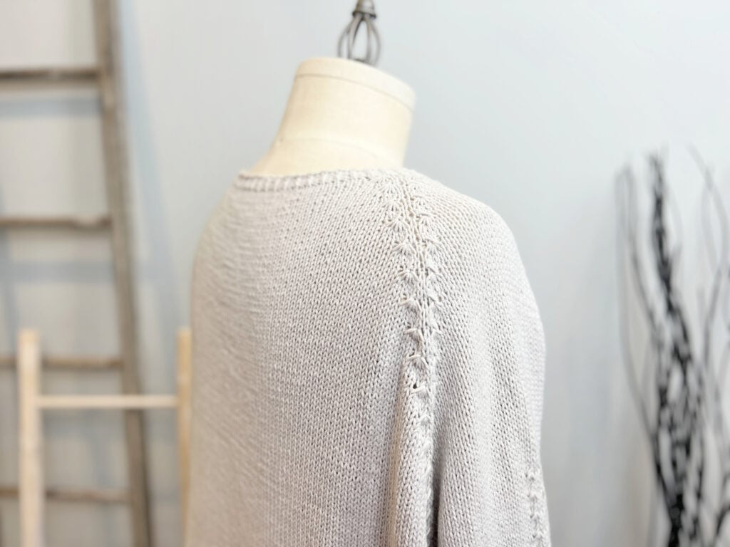 A close-up of a light gray knit cardigan pattern displayed on a mannequin, showing detailed textured stitching along the shoulder seam. The softly blurred background features a ladder and decorative branches, perfect for Marly Bird fans. -Marly Bird
