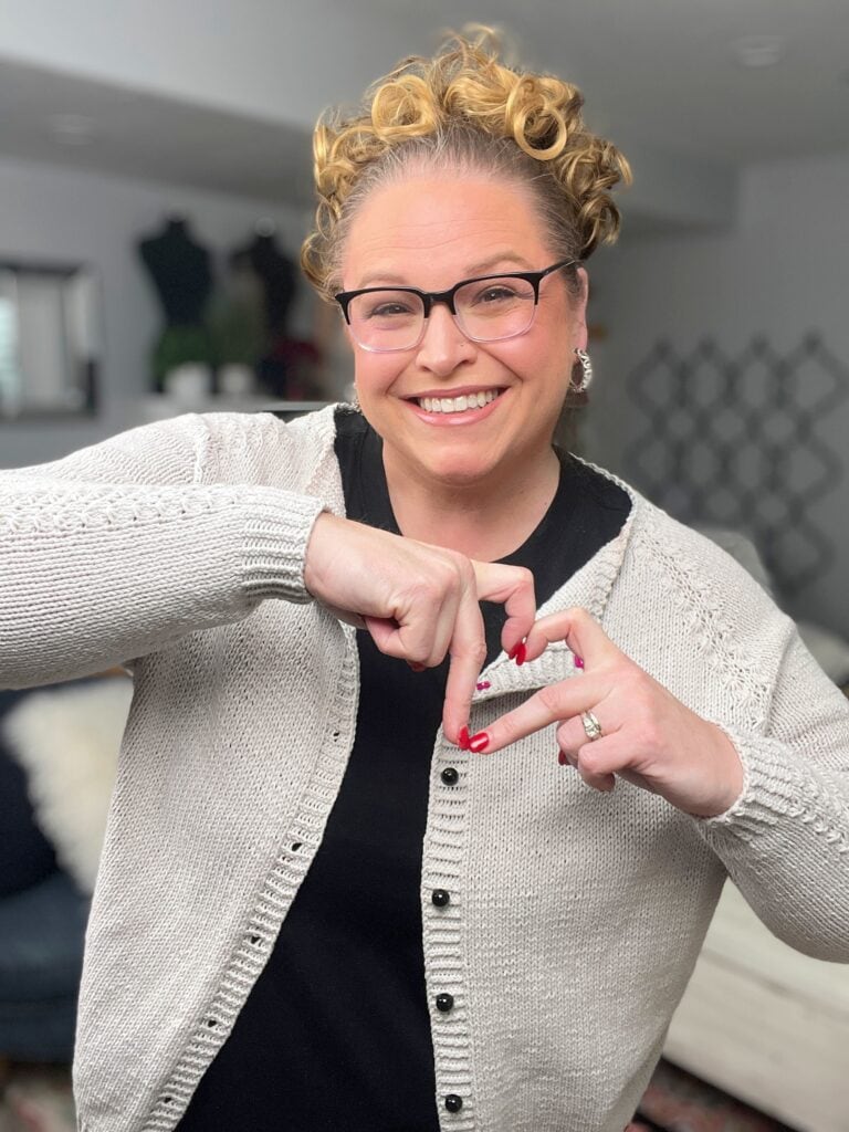 A smiling person with curly blonde hair and glasses makes a heart shape with their hands. They wear a light-colored cardigan—perhaps inspired by free knitting and crochet patterns—standing in a cozy, well-lit room with modern decor. -Marly Bird