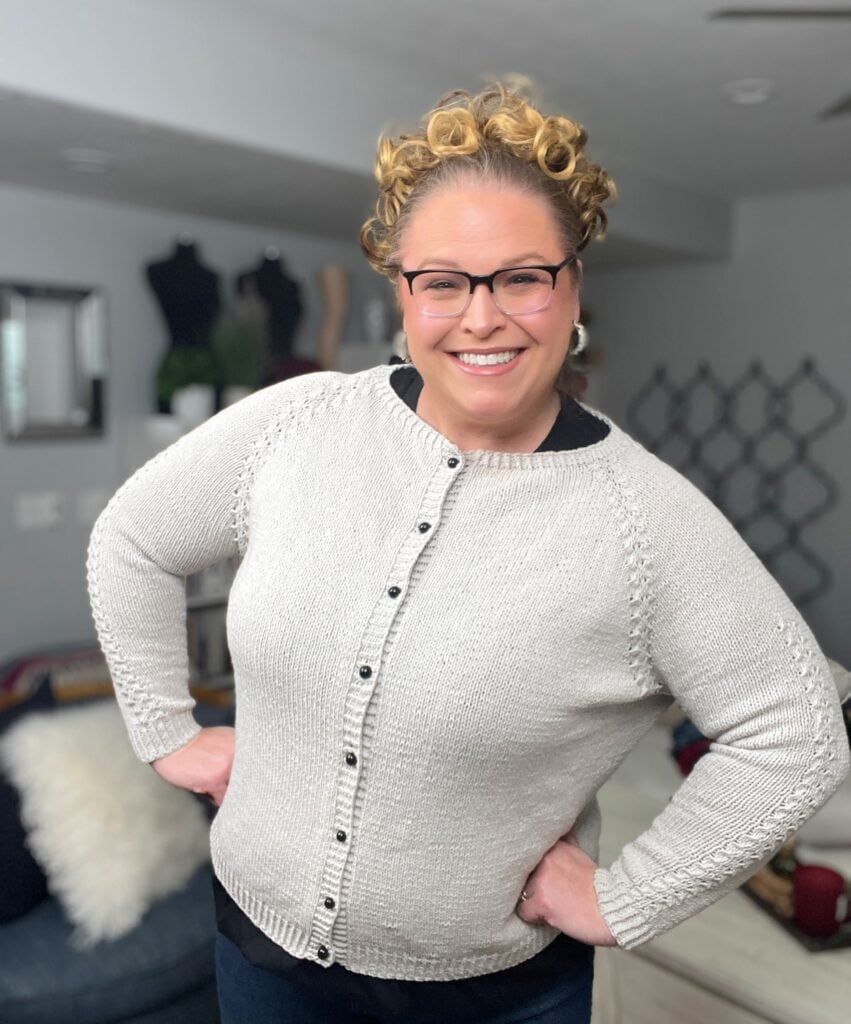A smiling person with curly hair and glasses wears a light-colored, buttoned knit sweater—perhaps crafted from free knitting and crochet patterns—and stands indoors with hands on hips; the background features home decor and mannequins. -Marly Bird