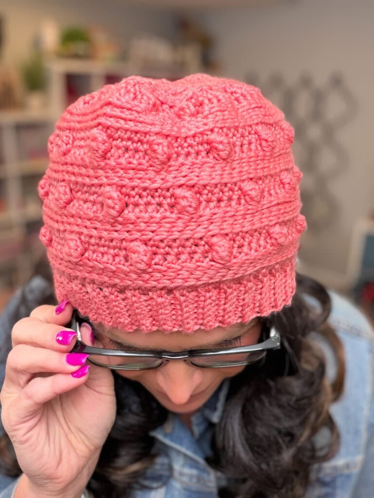 Close-up overhead view of Marly Bird peering over her glasses, showing the top and body of the Summit Chic crochet hat and the raised three-dimensional bobble stitch pattern across the crown. -Marly Bird