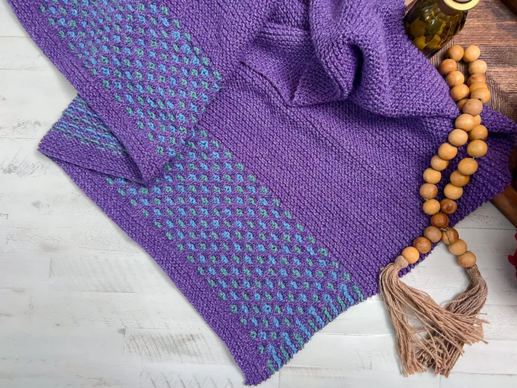A purple knitted scarf with blue and purple patterned details displayed on a white wood surface, showing stitch texture. Summit Knit Shawl free garter stitch knit shawl pattern