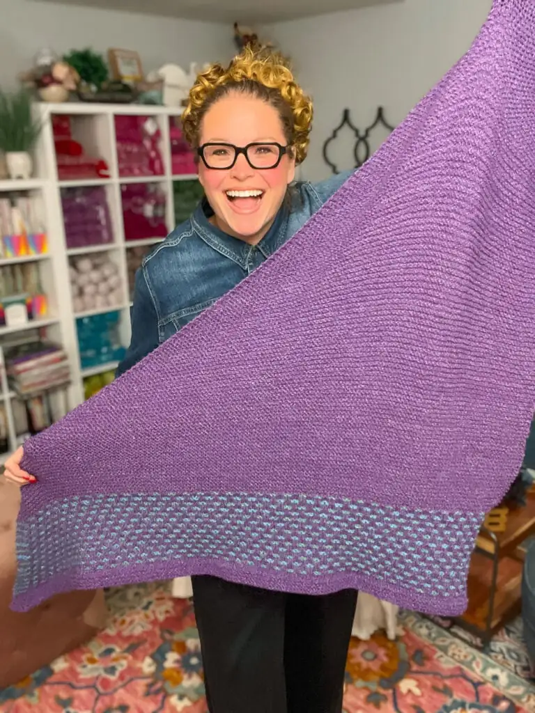 Person with curly hair models the Summit Knit Shawl, showing off its vibrant colors and textured knit stitches in a craft room. Summit Knit Shawl free garter stitch knit shawl pattern