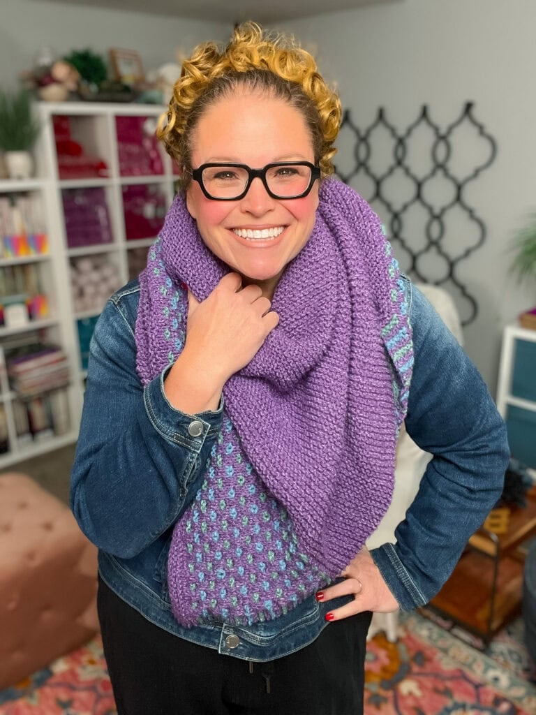 Person wearing a chunky, purple knit shawl with textured stitches over a denim jacket in a yarn-filled craft room.