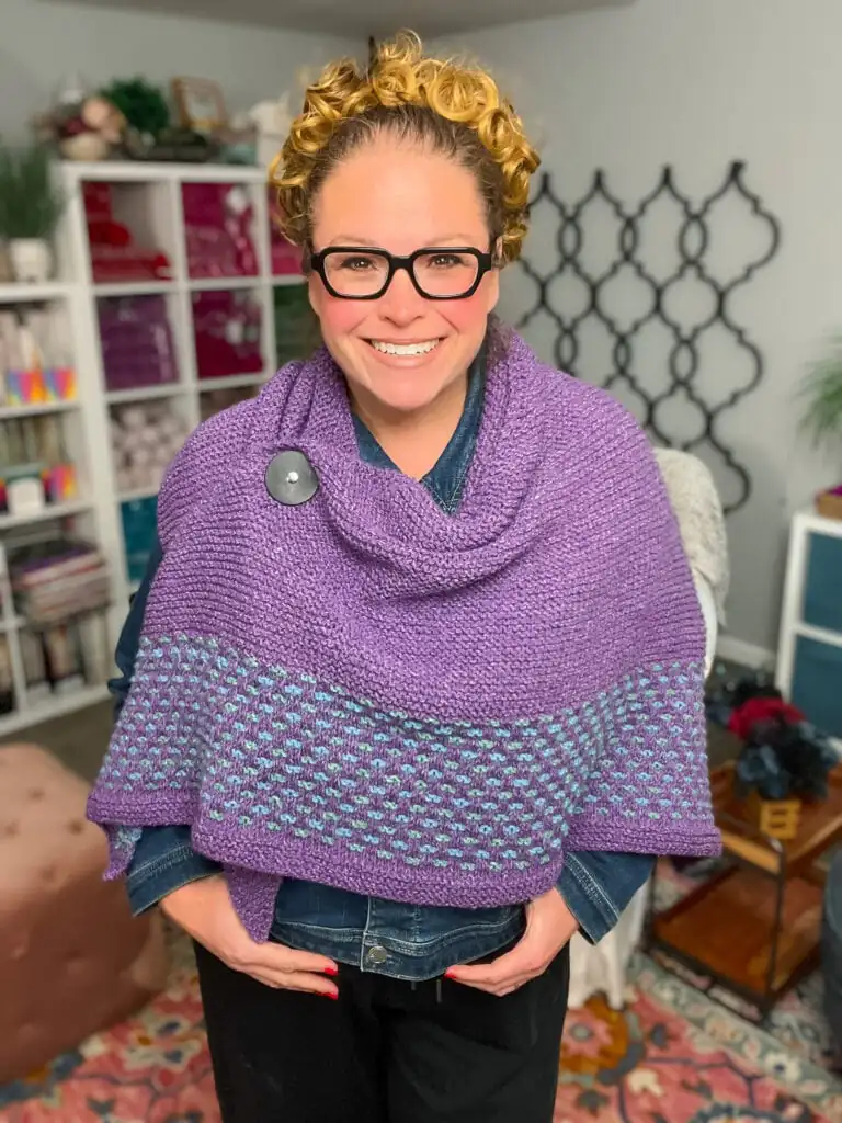 Woman models a purple knit shawl with a button closure and patterned trim; yarn and books are visible on shelves behind her. Summit Knit Shawl free garter stitch knit shawl pattern