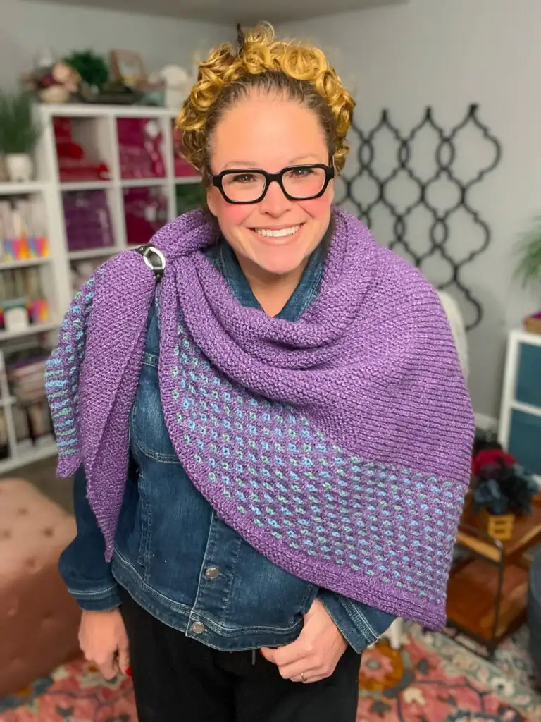 Woman models a hand-knit purple shawl with textured stitches over a denim jacket; yarn and plants fill shelves behind her. Summit Knit Shawl free garter stitch knit shawl pattern