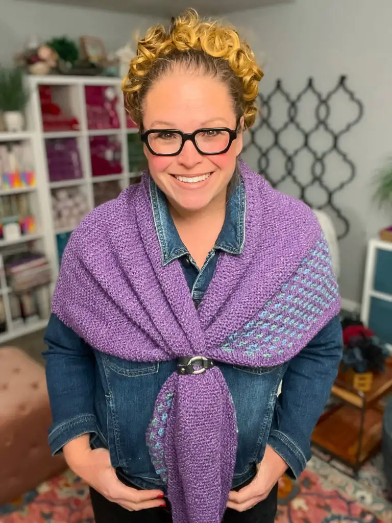 Person wears a textured purple knit shawl over a denim jacket; yarn and crafts visible on shelves in the cozy room behind. Summit Knit Shawl free garter stitch knit shawl pattern