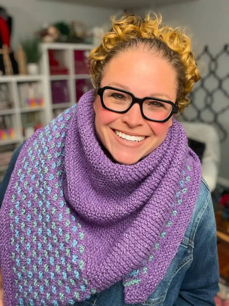 Purple Summit Knit Shawl with teal accents drapes over a denim jacket, highlighting intricate knit texture indoors near yarn shelves. Summit Knit Shawl free garter stitch knit shawl pattern
