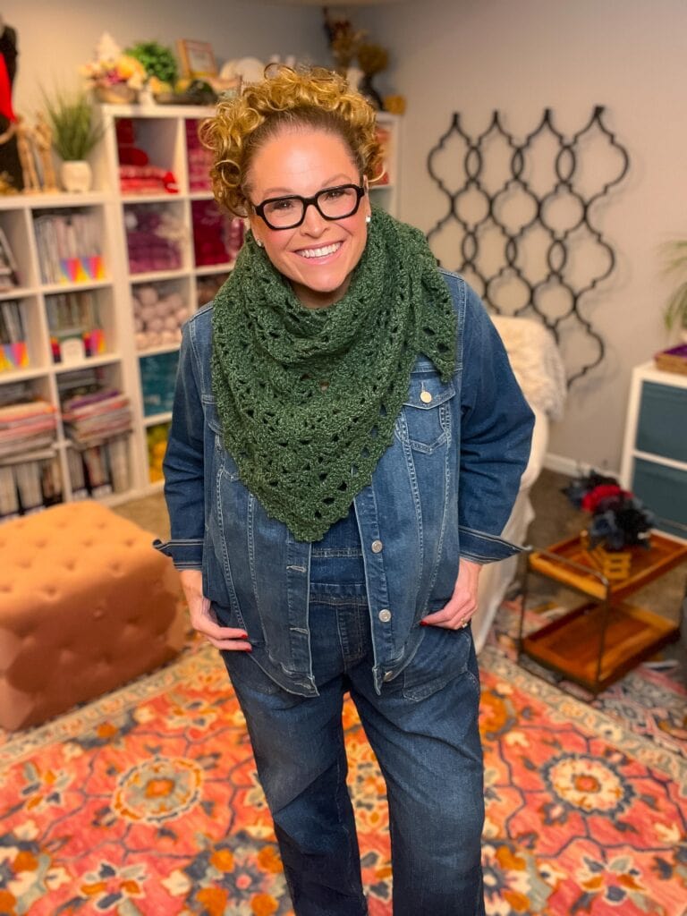 Person models a green crocheted scarf with visible textured stitches, standing in a craft room with yarn and bookshelves.