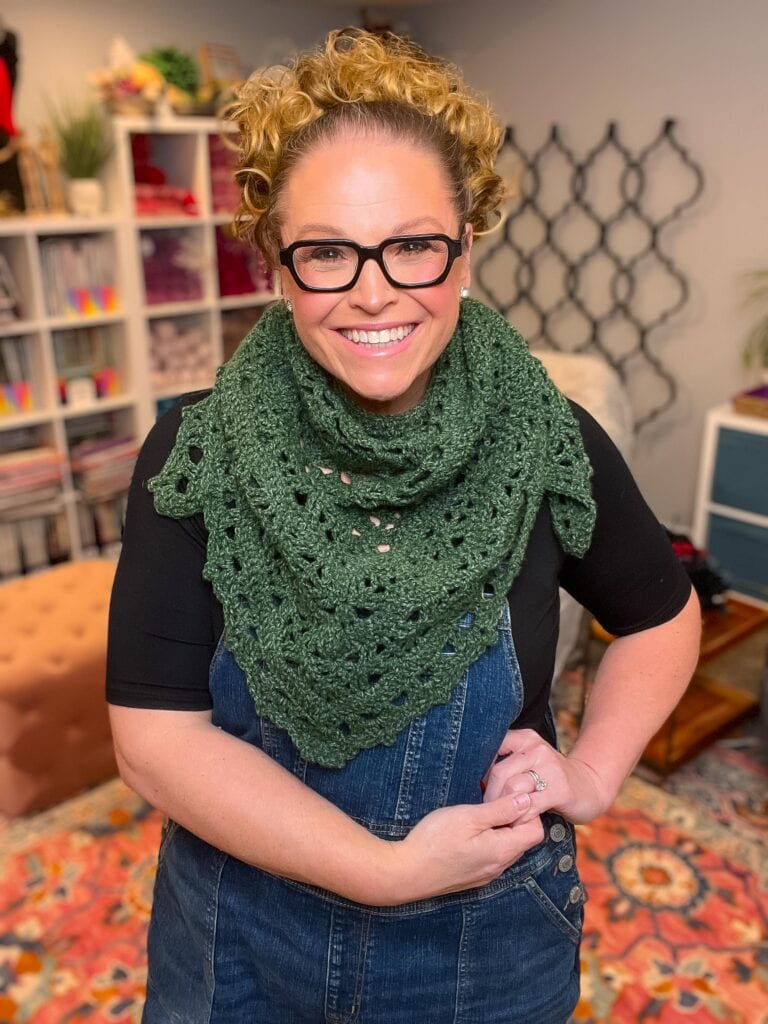 Person modeling a green crocheted shawl with visible beginner-friendly stitches, worn over overalls indoors.