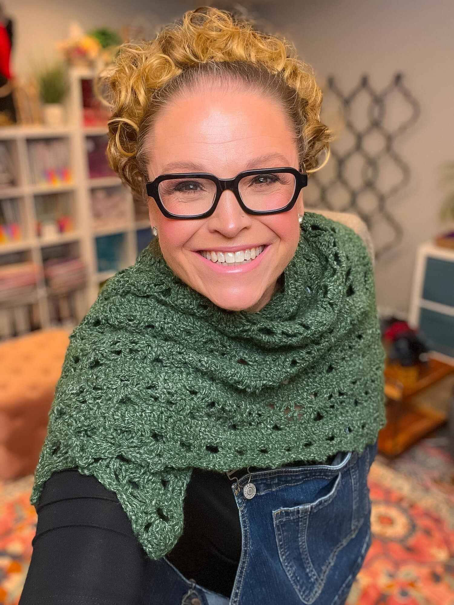 Person models a green crochet shawl with visible stitch detail, layered over black shirt and overalls in a cozy room.