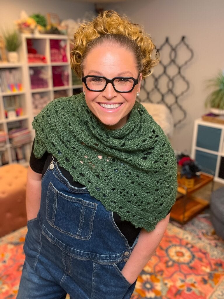 Green crocheted beginner shawl with visible stitch texture, modeled in a colorful room with books, yarn, and plants behind.
