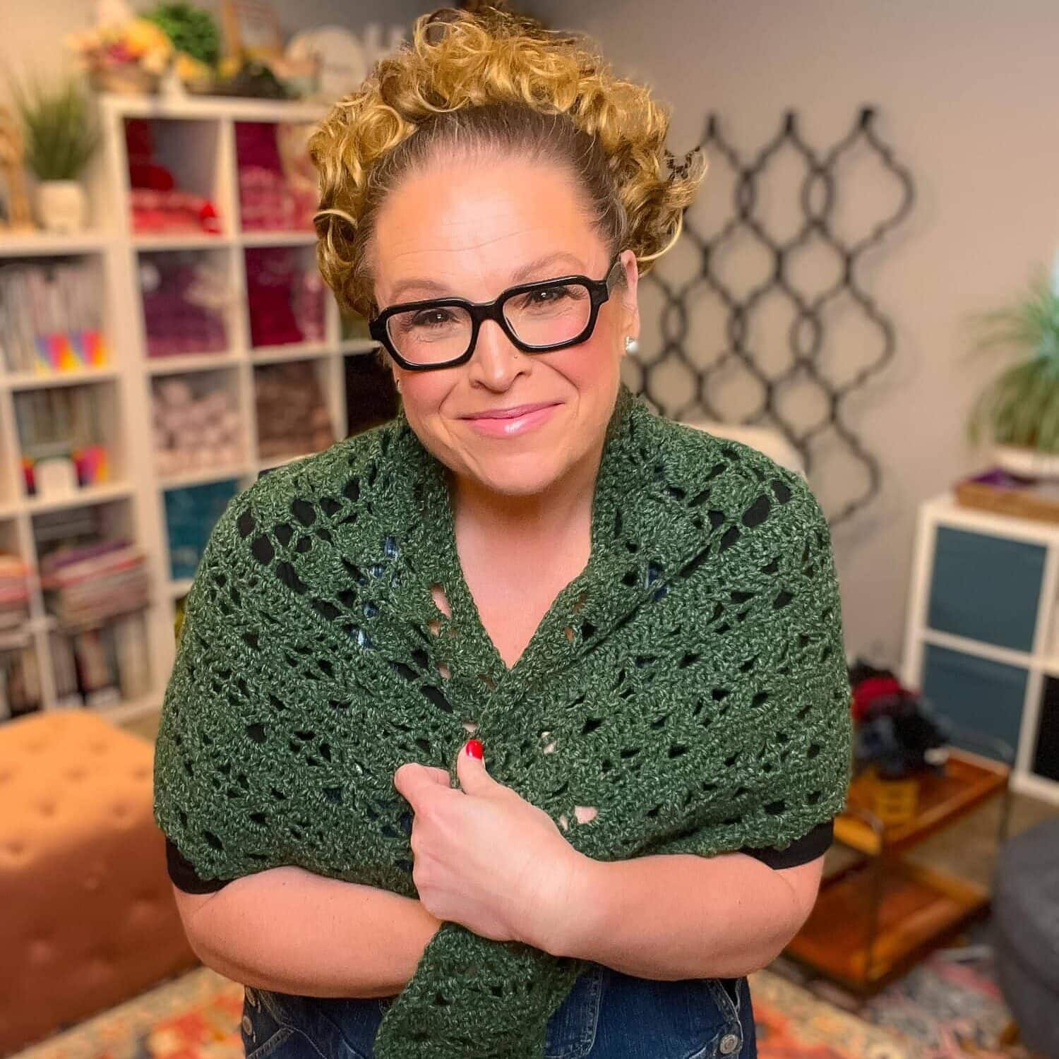 A green crochet shawl in worsted weight yarn, draped on a woman, shows textured stitches; cozy room background.