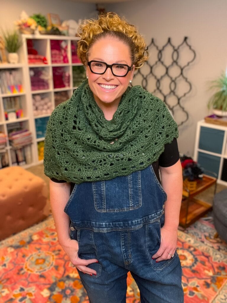 Person models a green worsted crochet shawl with simple stitches and soft drape, styled over denim in a bright, yarn-filled room.