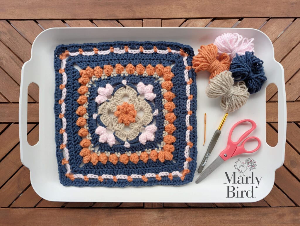 A Sweetcorn Motif crochet block in blue, orange, beige, and pink lies on a white tray beside yarn, crochet hooks, and pink scissors. The Marly Bird logo is in the bottom right corner. -Marly Bird