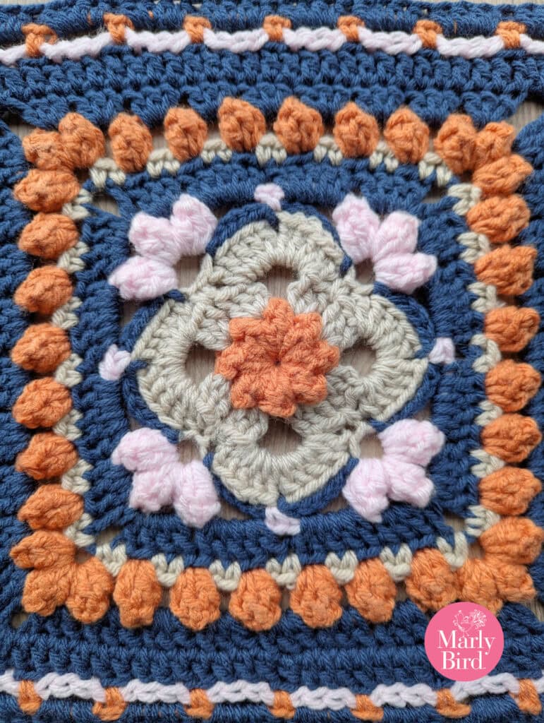A crocheted square featuring blue, orange, beige, and light pink yarn in a geometric floral pattern, inspired by the Sweetcorn Motif crochet block, with the "Marly Bird" logo in the bottom right corner. -Marly Bird