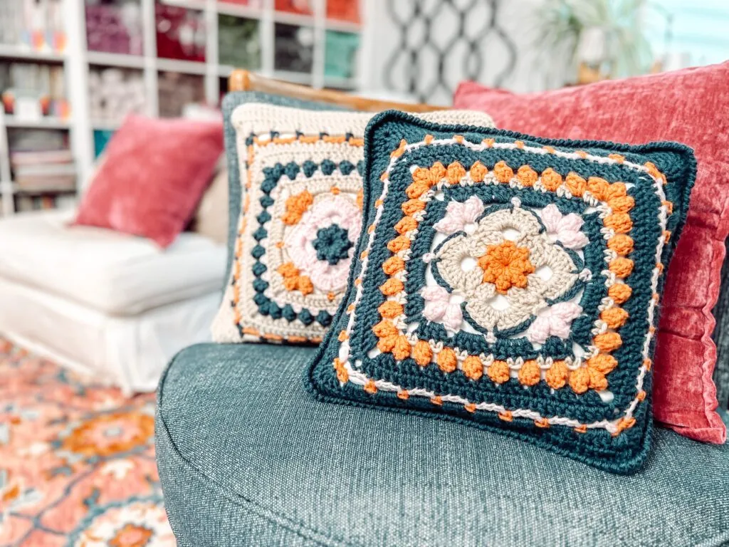 Two crochet pillows with colorful, geometric granny square and Sweetcorn Motif crochet block patterns rest on a blue sofa in a cozy living room with a red rug and blurred shelves in the background. -Marly Bird