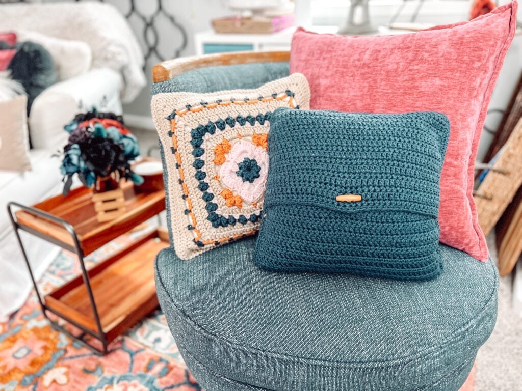 A teal chair displays three pillows: one pink, one teal with a wooden button, and one crocheted in a Sweetcorn Motif crochet block with cream, orange, pink, and teal. A cozy living room setup is visible in the background. -Marly Bird