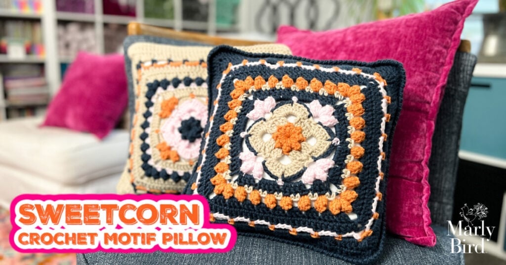 Two colorful crochet motif pillows with geometric patterns in navy, beige, orange, and pink add a pop of crochet home décor to a couch with bright pink cushions. Text reads "Sweetcorn Crochet Motif Pillow" with the Marly Bird logo in the corner. -Marly Bird