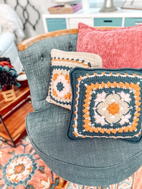 Two decorative pillows with colorful crocheted granny square designs rest on a blue upholstered chair, showcasing the charm of free knitting and crochet patterns. One pillow is pink, while the other features green, orange, and cream crochet motifs. -Marly Bird