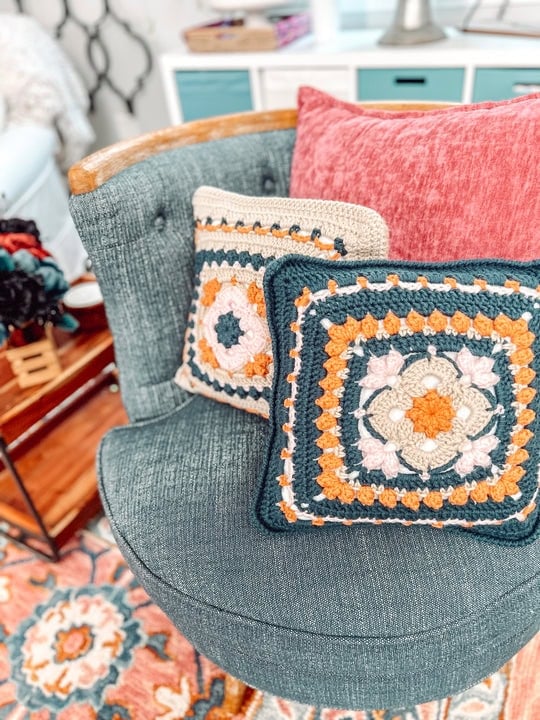 A blue chair with two colorful crocheted pillows—crafted using free knitting and crochet patterns—featuring geometric designs sits by a pink pillow. In the background, a floral rug and a wooden side table with yarn complete the cozy scene. -Marly Bird