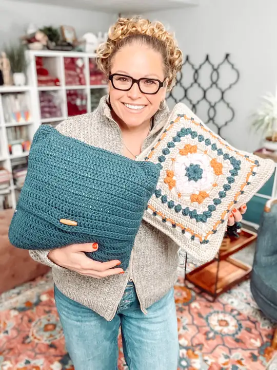 A smiling woman with curly hair and glasses holds two crocheted pillows, one solid blue with a button and the other with a colorful granny square pattern. She stands in a cozy living room, inspired by free knitting and crochet patterns. -Marly Bird