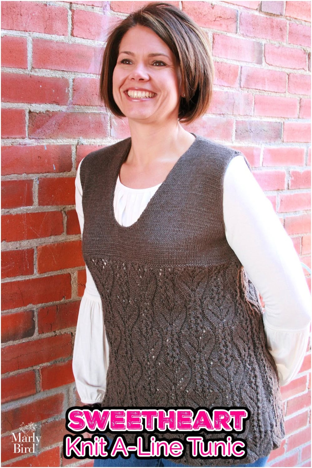 A smiling woman stands against a red brick wall, wearing a white long-sleeve shirt under a brown, knit A-Line Tunic with a lace pattern. Pink text at the bottom reads, "SWEETHEART Knit A-Line Tunic by Marly Bird. -Marly Bird