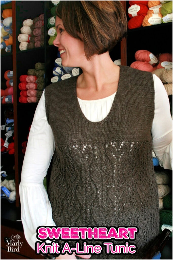 A woman wearing a brown, sleeveless knit A-Line Tunic with a lace pattern stands in front of shelves filled with colorful yarn. Text reads: "SWEETHEART Knit A-Line Tunic—Free Knit Tunic Pattern by Marly Bird. -Marly Bird
