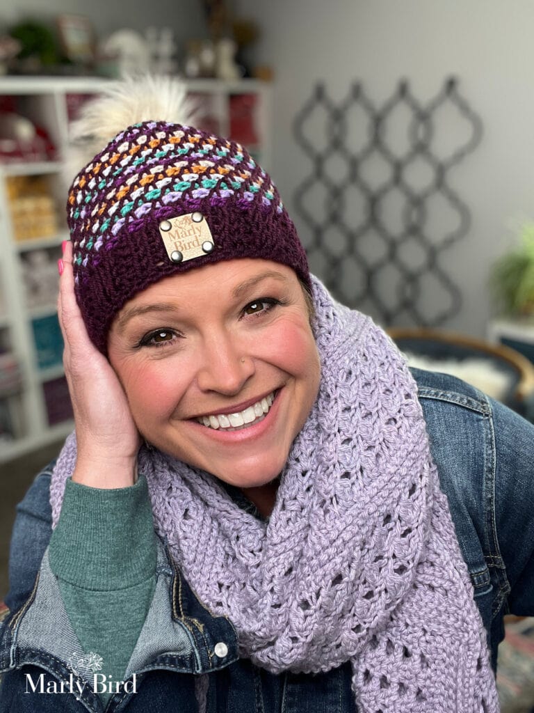 A smiling woman wears a colorful knit beanie with a "Marly Bird" tag and a lavender crocheted scarf—perhaps inspired by a free hat pattern. She rests her hand on her face, standing in a cozy, yarn-filled room. -Marly Bird
