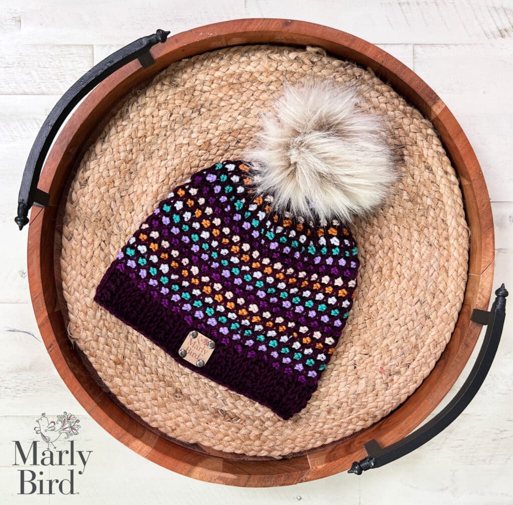 A multicolored crocheted beanie made with Tunisian crochet and topped with a large faux fur pom-pom sits on a woven tray. The beanie features purple, orange, blue, and white stitches with a dark ribbed brim and small tag. “Marly Bird” appears below. -Marly Bird
