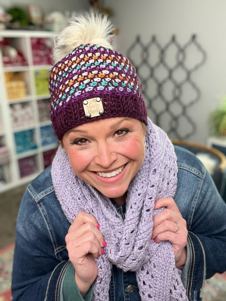 A smiling woman wearing a colorful knit beanie with a pom-pom, made from a free hat pattern, and a light purple crochet scarf. She is dressed in a denim jacket, standing indoors with yarn shelves and a patterned wall in the background. -Marly Bird
