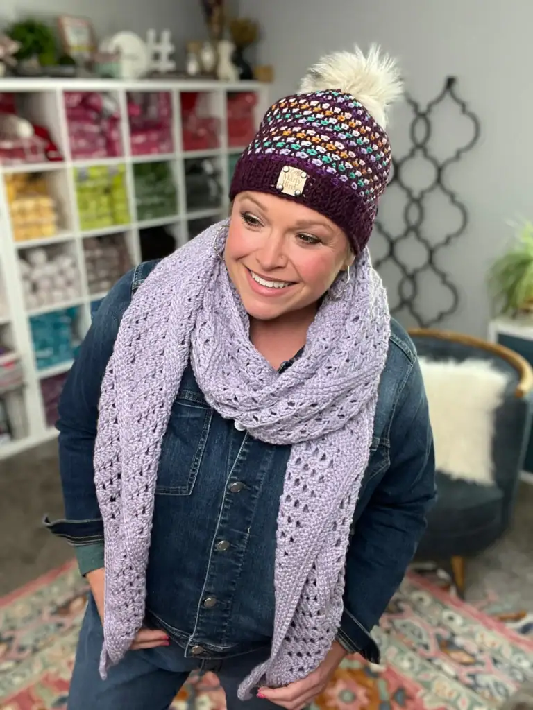 A smiling woman in a denim jacket and colorful knit hat wears a chunky lavender crochet scarf. She stands indoors in front of a yarn-filled shelf, perhaps searching for free knitting and crochet patterns to inspire her next creation. -Marly Bird