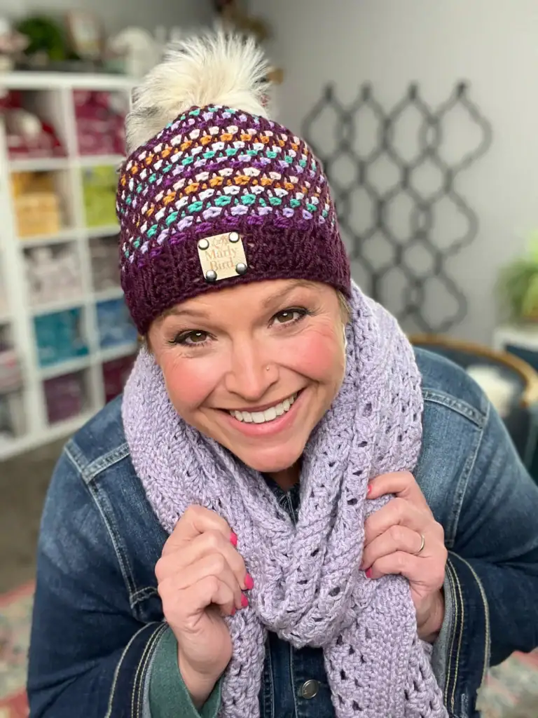 A smiling woman in a multicolored knit hat with a pom-pom and “Old Mary Hero” tag, plus a lavender crochet scarf and denim jacket, stands indoors by shelves of yarn—showcasing her love for free knitting and crochet patterns. -Marly Bird